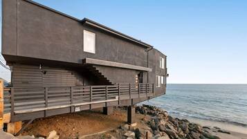 A modern coastal house with a balcony, built on a rocky shoreline.