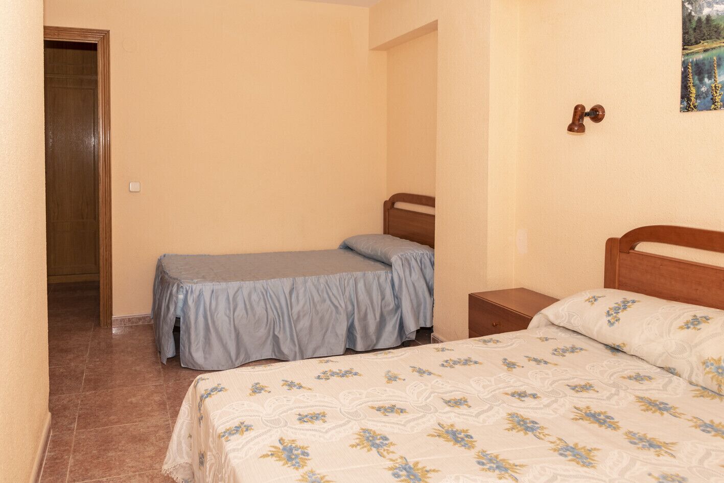A small, simple bedroom with two single beds, a wooden headboard, a nightstand, and a door leading to another room.