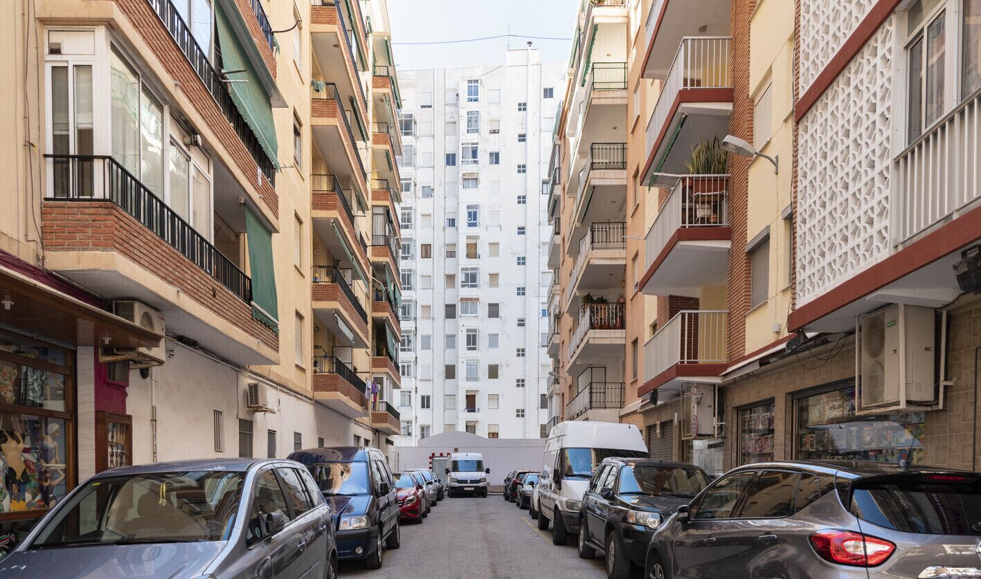 Una calle estrecha en la ciudad, bordeada por edificios residenciales de varios pisos con balcones, coches aparcados y una ambulancia visible en la distancia.