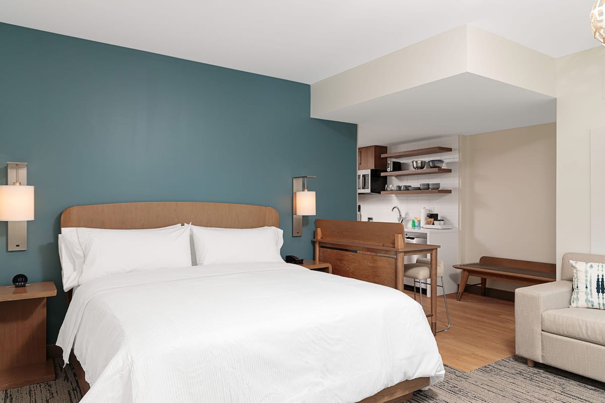 Een hotelkamer met een groot bed, nachtkastjes, een bureau, een stoel, een bank en een kitchenette op de achtergrond.