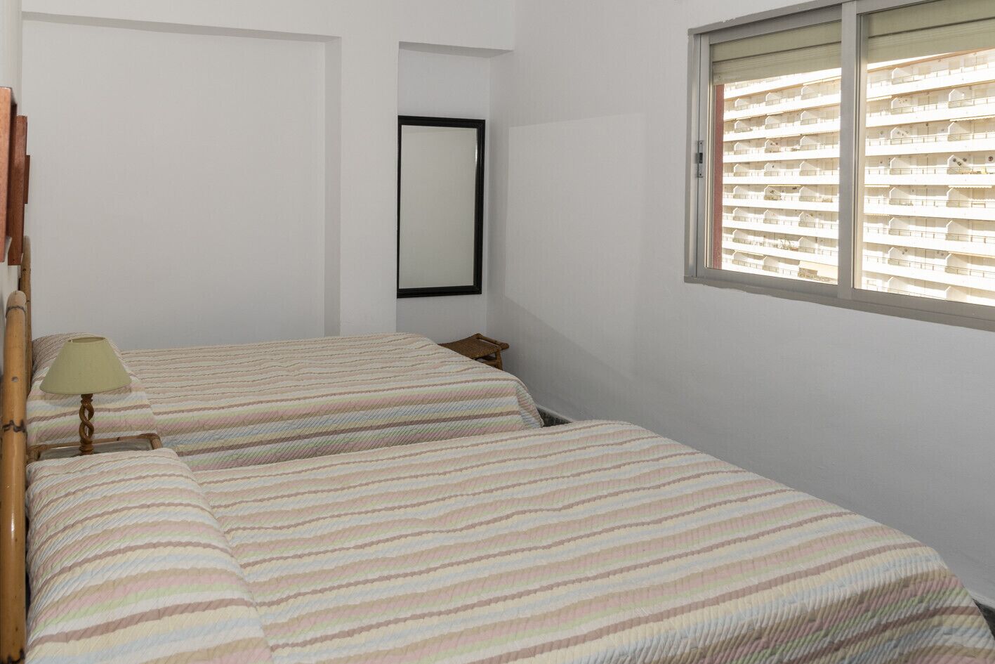 Une petite chambre propre, avec deux lits, une table de chevet, une lampe et une fenêtre avec des stores.