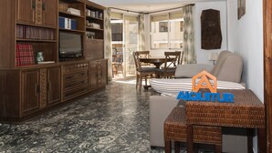 A living room with a sofa, a television, and a bookshelf. There is a dining table with chairs and a window with curtains. - EL RACO, C-8º-16 (Cullera)