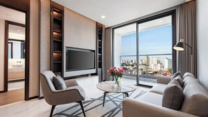 Un salón moderno con un televisor de pantalla plana grande, asientos cómodos, una mesa de centro y vistas a un paisaje urbano a través de amplias ventanas.