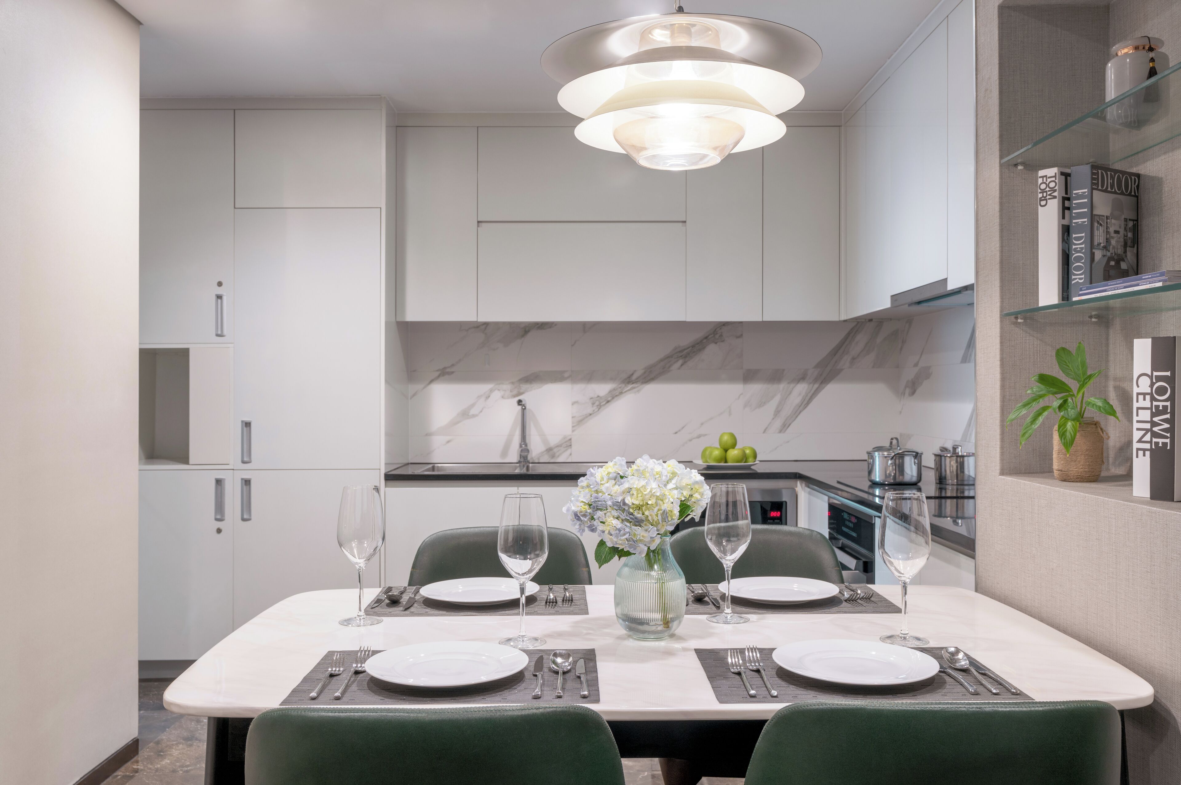 A modern dining area with a marble countertop, white cabinets, and a glass shelf with books and a plant.