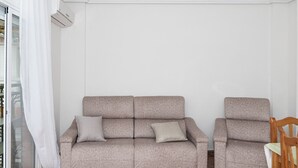 Ein Wohnzimmer mit grauem Sofa und einem Holzstuhl, eine Glastür mit weißem Vorhang und einer weißen Wand mit Klimaanlage.