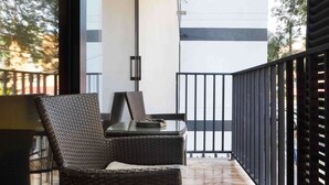 Varanda com mobiliário de vime e mesa de vidro, com vista para um edifício com fachada branca e guarda-corpo preto.