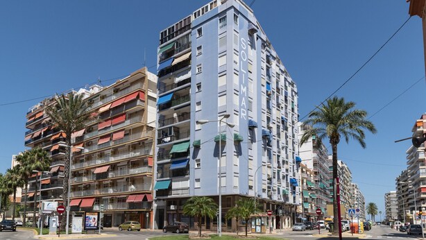 Un edifici de diversos pisos amb balcons, un carrer amb palmeres i un cel blau clar.