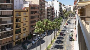 Un edifici d'apartaments de diverses plantes amb balcons, un carrer amb palmeres i cotxes aparcats.