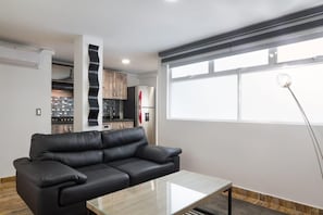 Un salon moderne avec un canapé en cuir noir, une table basse en verre et un coin cuisine doté de meubles en bois et d’un réfrigérateur.