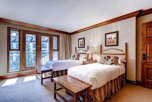 Une chambre d’hôtel avec deux lits, un banc en bois, un bureau et de grandes fenêtres donnant sur des immeubles.