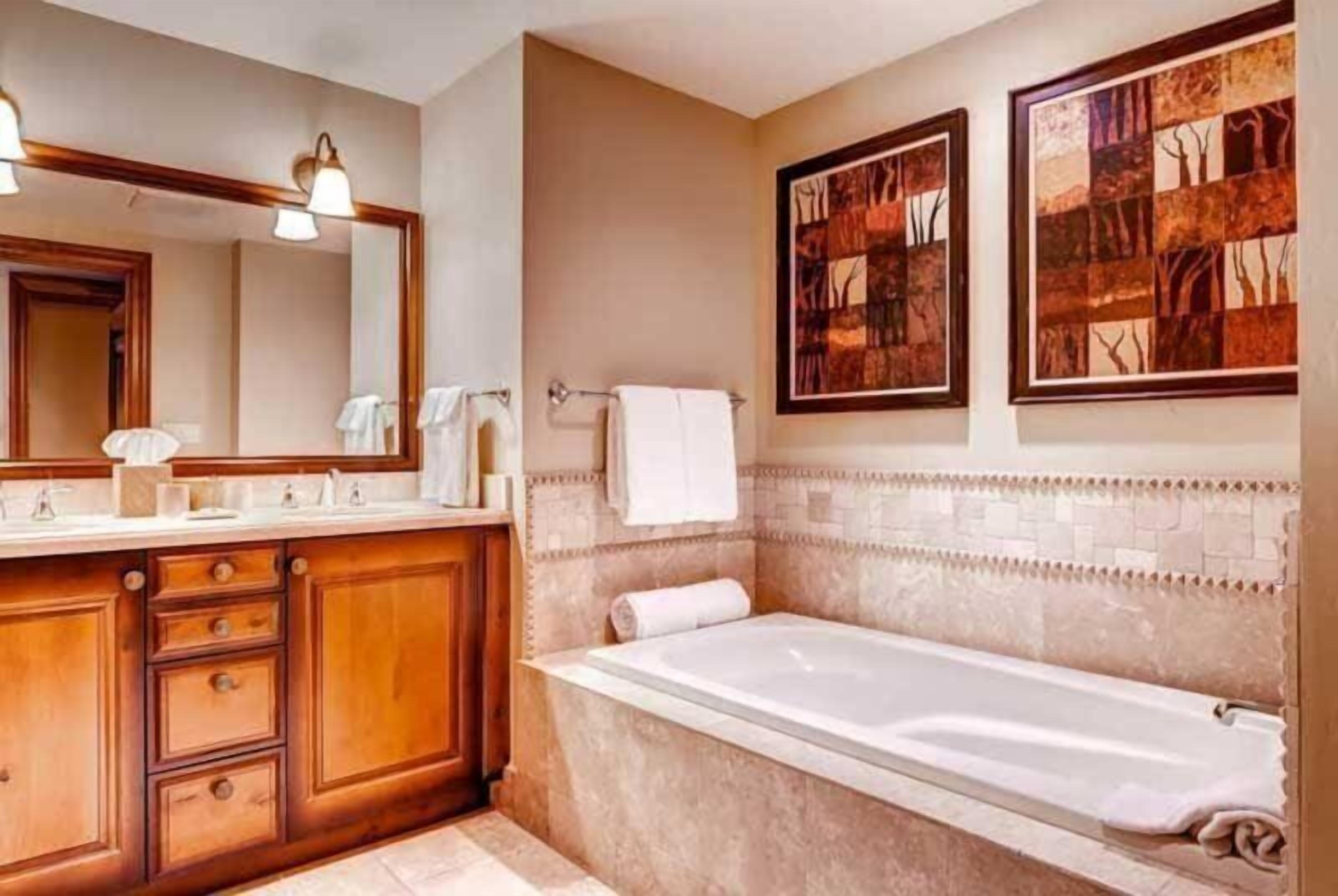 A bathroom with a wooden vanity, a large mirror, a bathtub, and two framed artworks on the wall.