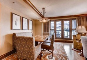 A dining area with a wooden table and patterned chairs, a chandelier, and a view of a balcony with curtains. - Beaver Creek Park Hyatt 3 Bedroom Residence (Avon)