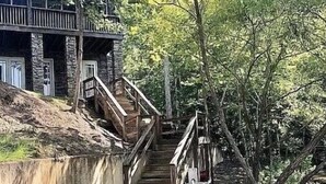 Una scala in legno scende verso la riva di un fiume, circondata da alberi e un edificio con facciata in pietra.