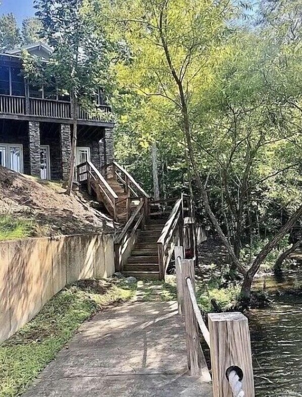 A wooden staircase leading down to a riverbank, surrounded by trees and a building with a stone facade. - Waterfront house, Minutes to Auburn, Boat slip. (Dadeville)