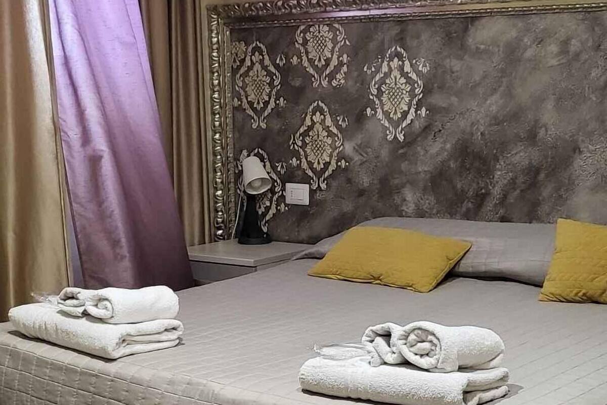 A hotel room with a bed, pillows, a bedside table, an air conditioner, and curtains.