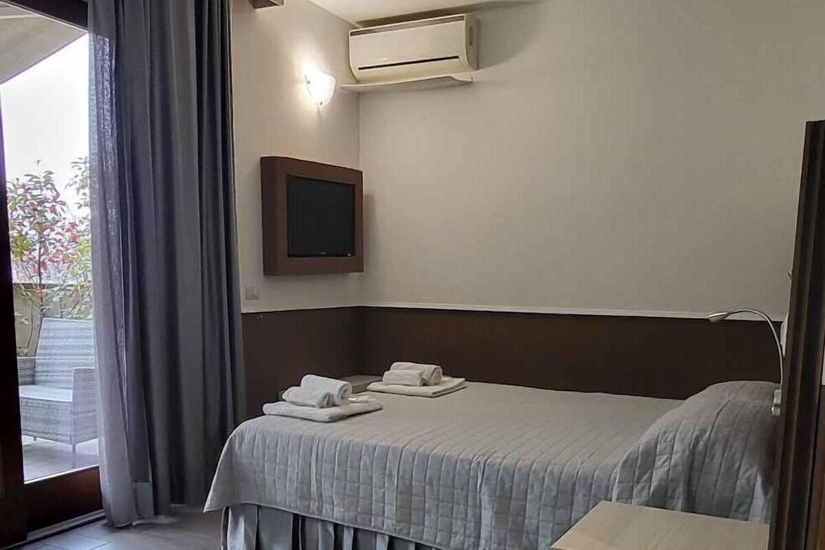 A hotel room with a bed, a TV, an air conditioning unit, and a view of the outdoors.