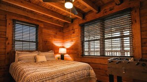 Un dormitorio de cabaña de madera con una cama, dos ventanas con persianas, una mesita de noche con una lámpara y un aparador de madera.