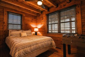 A wooden cabin bedroom with a bed, two windows with blinds, a nightstand with a lamp, and a wooden chest of drawers.
