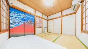 Une pièce de style japonais traditionnel, avec un sol recouvert de tatamis, un lit, une porte coulissante ornée d’une peinture de montagne et un ventilateur de plafond.