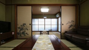 A traditional Japanese room with tatami flooring, a low table with a decorative mat, sliding doors with shoji screens, and a television on a wooden stand. - Hikari no Yado - Detached house for rent in Anan City, Tokushima Prefecture (徳島県)