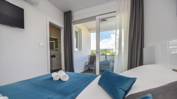 A hotel room with a bed, a blue blanket, a towel, a TV, a sliding glass door to a balcony, and a small table with a chair. - Mare Room with Balcony and Shared Pool (Novalja)