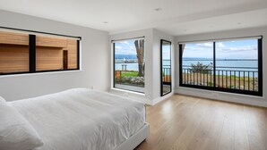 Een moderne slaapkamer met een groot bed, houten vloeren en grote ramen met uitzicht op zee en een brug.