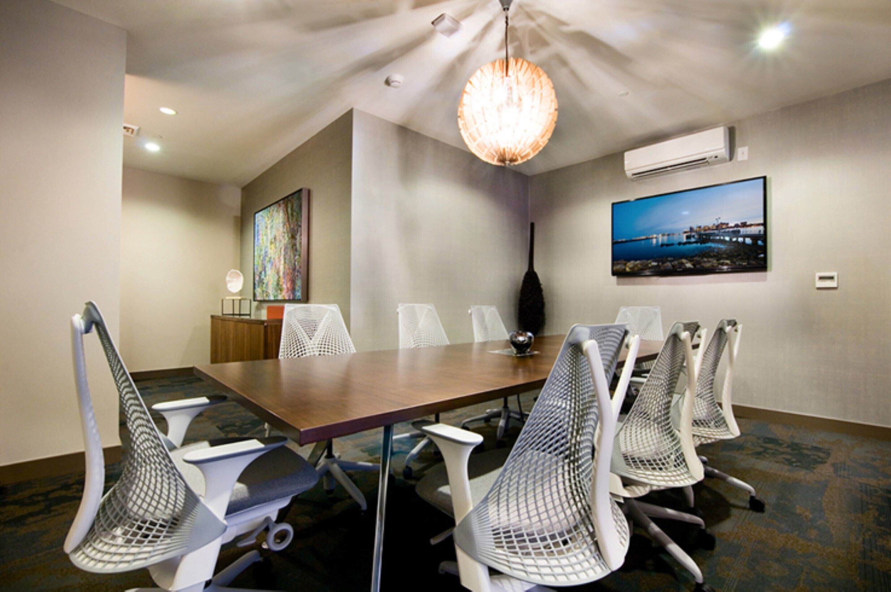A conference room with a long table, six mesh chairs, a chandelier, an air conditioning unit, and a painting on the wall.