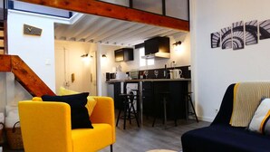 A modern living room with a yellow armchair, a dark sofa, and a wooden coffee table. A kitchen is visible in the background with a bar area. - 1 Bedroom with terrace - Wifi, Air Conditionning / 1 to 5 People (Aix-en-Provence)