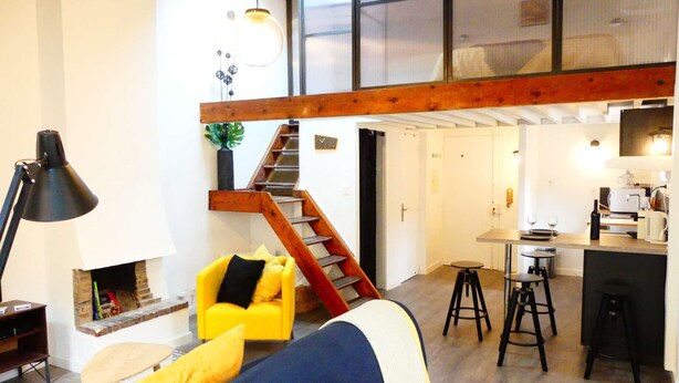 A spacious living area with a high ceiling, a wooden staircase, a kitchen with stools, and a cozy seating area with a yellow armchair. - 1 Bedroom with terrace - Wifi, Air Conditionning / 1 to 5 People (Aix-en-Provence)