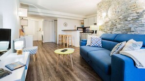 A modern living room with a blue sofa, a small round table, and a TV on a white stand. There is a kitchen in the background with a stone wall. - 1 Bedroom Flat Quartier Rotonde - Lift, Wifi, Air Conditionning / 1 to 6 People (Aix-en-Provence)