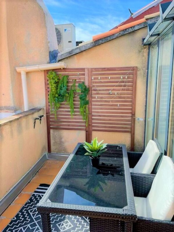 Une petite terrasse extérieure avec une table à plateau de verre et des chaises blanches rembourrées, un pot de fleurs et une clôture en bois ornée de verdure.