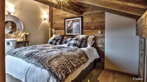 A cozy bedroom with a wooden ceiling, a bed with multiple pillows, a round mirror, and a framed picture on the wall.