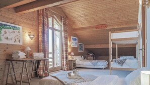 Ein gemütliches Blockhauszimmer mit Holzwänden, einem Schlafplatz auf einer Erhöhung, einem Schreibtisch mit Lampe und einem Fenster mit Vorhängen.