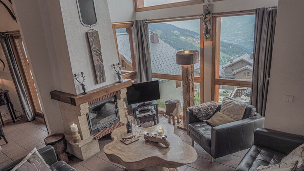 A spacious living room with a fireplace, leather sofas, a wooden coffee table, and a large window offering a view of the mountains.