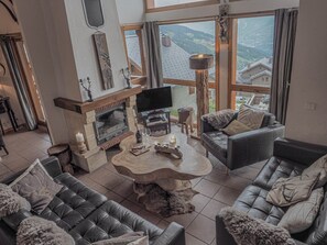 A spacious living room with a fireplace, leather sofas, a wooden coffee table, and a large window offering a view of the mountains.