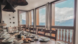 A dining area with a view of mountains through large windows, wooden chairs with heart-shaped backs, and a table set with plates, bowls, and a croissant. - Le Cairn, stunning 14 person, 7 bedroom chalet with sauna & games room (Peisey Nancroix)