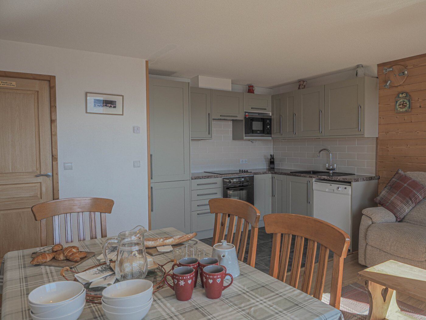 Uma cozinha acolhedora com uma mesa de jantar posta para quatro pessoas, com toalha de mesa xadrez, cadeiras de madeira e diversos itens para o café da manhã.