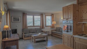 A cozy living room with a sofa, a coffee table, and a kitchen area with wooden cabinets and a stove. - Light, bright 2 bed apartment for 6 people with lovely views and close to lifts (Landry)