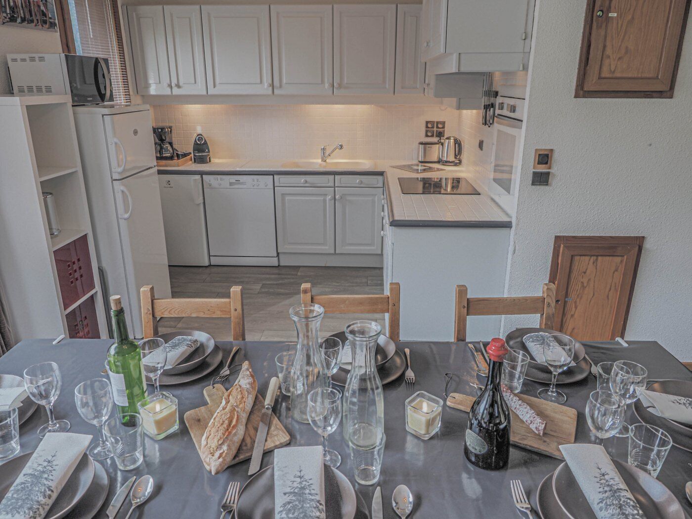 A modern kitchen with white cabinets, a dishwasher, and a dining area set for a meal with wine glasses, plates, and cutlery.