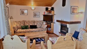 A cozy living room with a patterned sofa, wooden coffee table, and matching chairs. A fireplace and a TV stand with speakers are also visible.