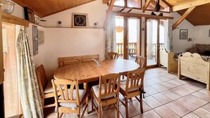 A wooden dining area with a round table and chairs, a clock on the wall, and a view of the outdoors through large windows.