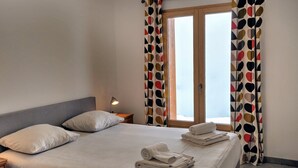 A neatly made bed with white linens and towels, a bedside lamp, and a window with curtains.