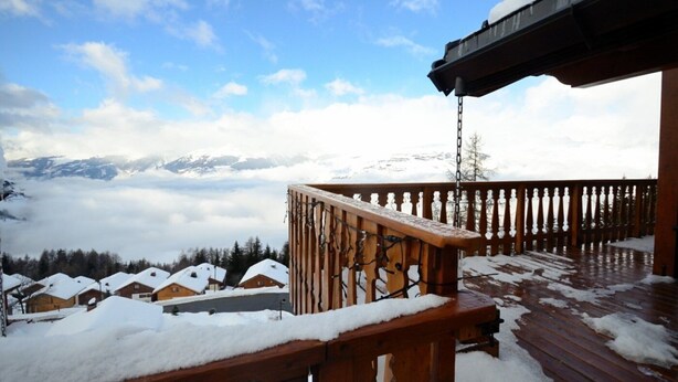 A wooden deck with snow-covered railings and a view of snow-covered houses and mountains in the background.