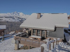 A snow-covered chalet with a balcony, wooden railings, and a view of snow-capped mountains.
