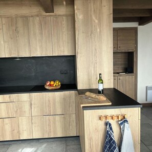 A modern kitchen with wooden cabinetry, a black countertop, a fruit bowl, a bottle of wine, and a cutting board.
