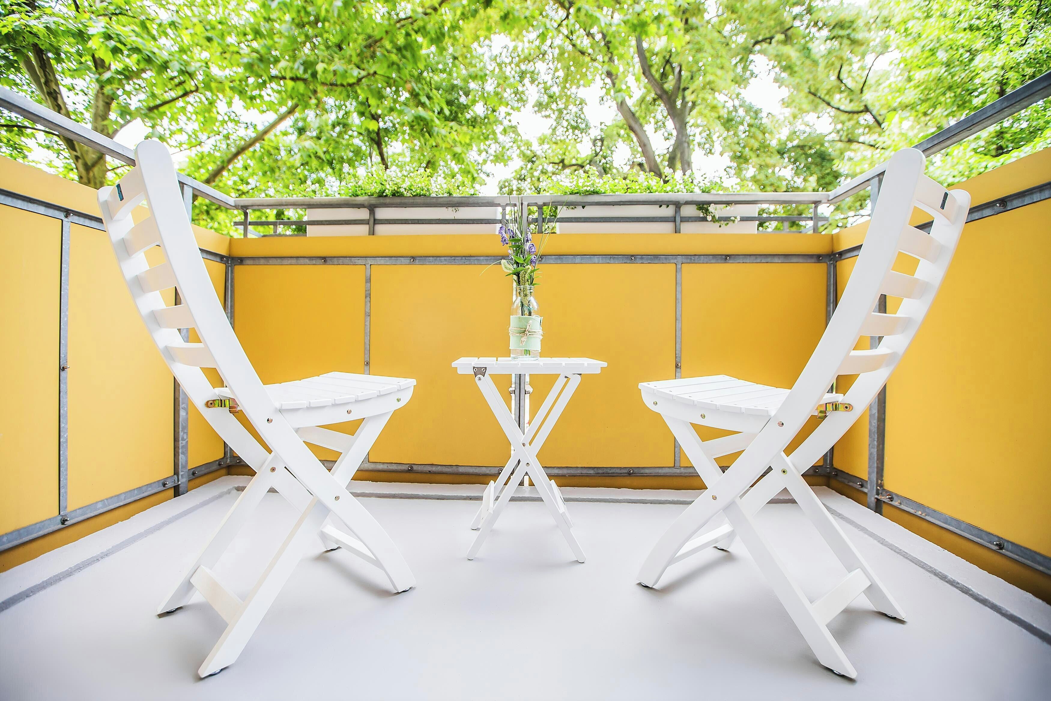 A balcony with two white chairs and a small table, yellow walls, and a view of green trees.