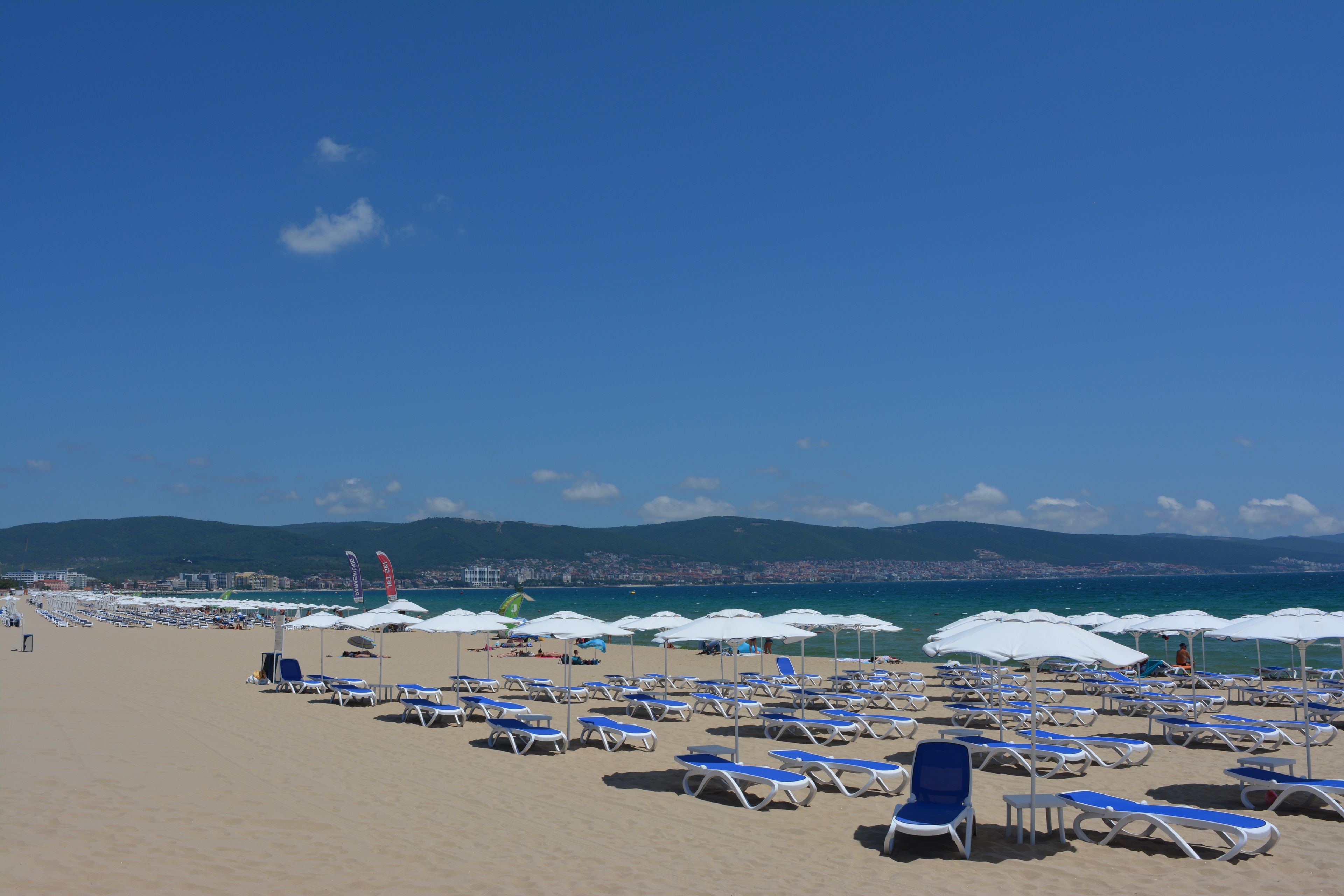 Ein Strand mit Reihen von Liegestühlen und Sonnenschirmen, einem klaren Himmel und fernen Bergen.