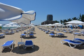 A beach with rows of sunbeds and umbrellas, a clear sky, and a building in the background. - Lotus (Nessebar)
