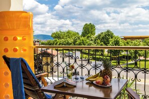 Un balcón con una mesa, una silla, una bebida, fruta y vistas a la calle y zonas verdes.