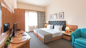 A hotel room with a bed, a desk with a TV, a chair, and a window with curtains. - Lotus (Nessebar)